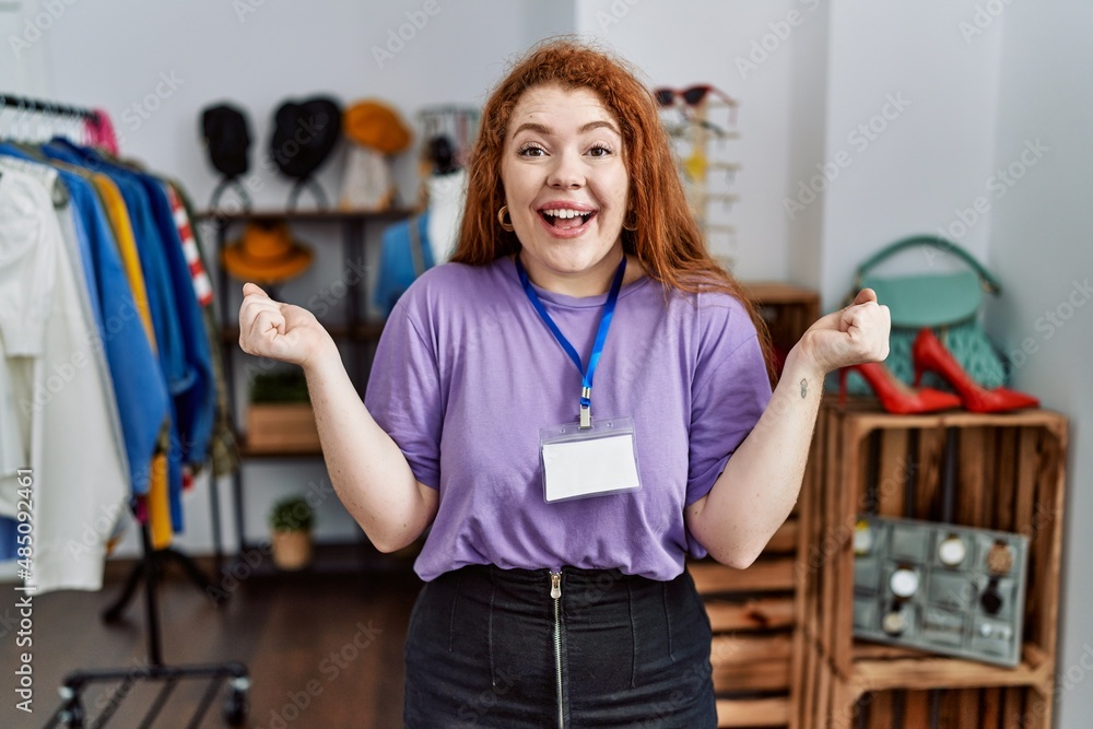 Young redhead woman working as manager at retail boutique celebrating surprised and amazed for success with arms raised and open eyes. winner concept. Young redhead woman working as manager at retail boutique celebrating surprised and amazed for success with arms raised and open eyes. winner concept.