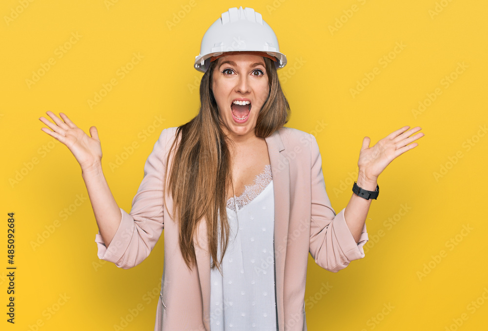 Young blonde woman wearing architect hardhat celebrating victory with happy smile and winner expression with raised hands Young blonde woman wearing architect hardhat celebrating victory with happy smile and winner expression with raised hands