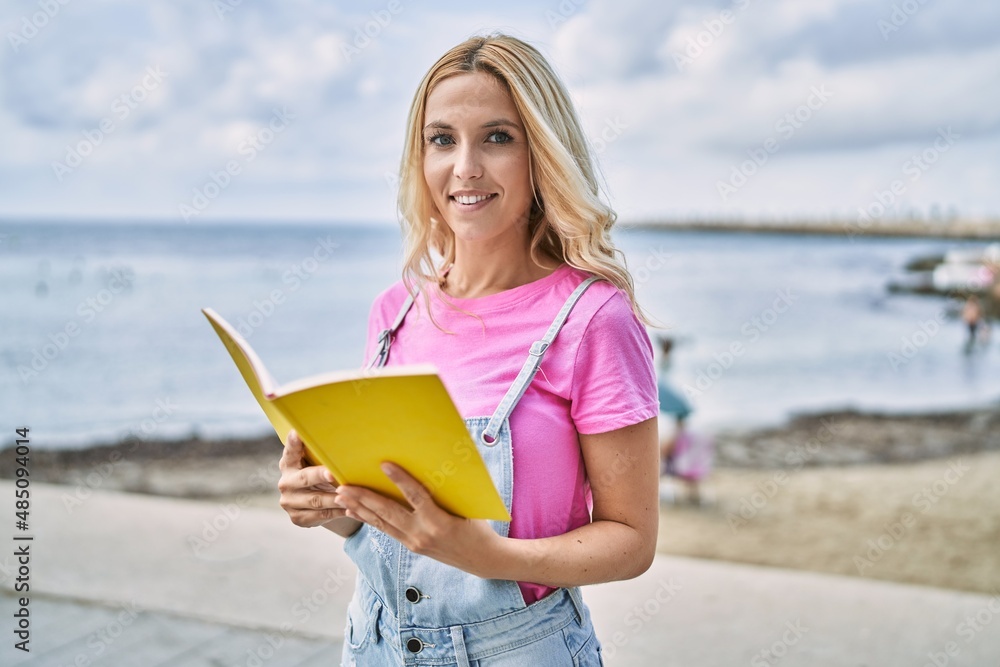 Young blonde woman smiling confident reading book at seaside