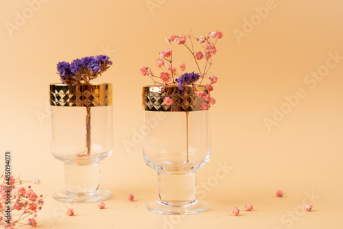Fototapeta Naklejka Na Ścianę i Meble -  Two beautiful glass goblets with golden rims and flowers inside on nude cream fabric