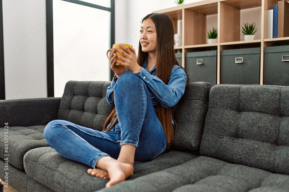 Obraz premium Young chinese girl drinking coffee sitting on the sofa at home.