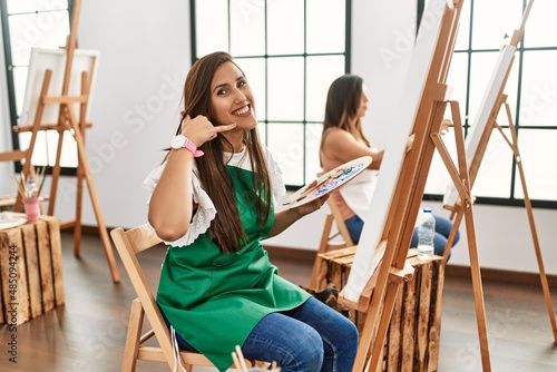 Young hispanic artist women painting on canvas at art studio pointing to the back behind with hand and thumbs up, smiling confident
