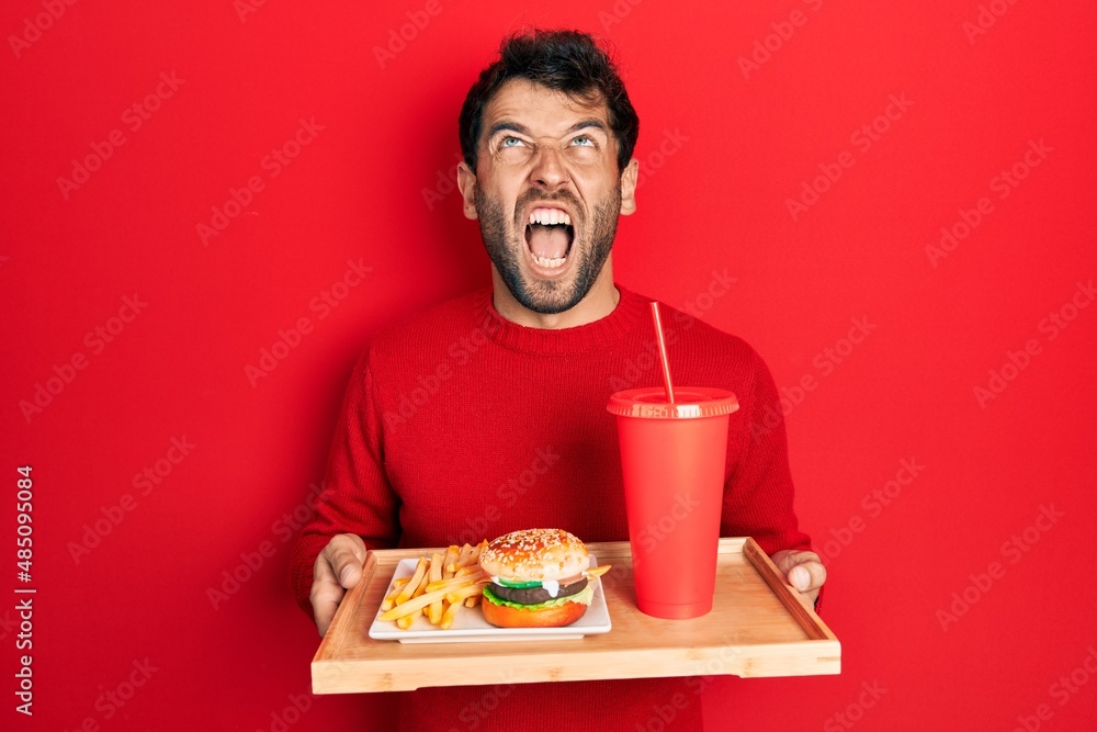 Handsome man with beard eating a tasty classic burger with fries and soda angry and mad screaming frustrated and furious, shouting with anger looking up. Handsome man with beard eating a tasty classic burger with fries and soda angry and mad screaming frustrated and furious, shouting with anger looking up.