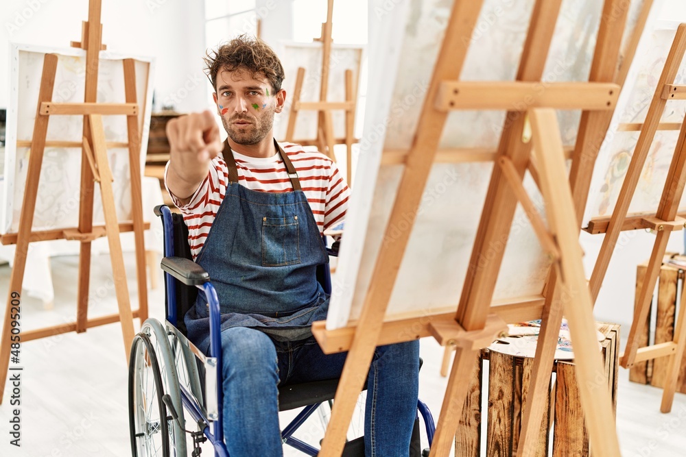 Young handsome man with beard at art studio sitting on wheelchair pointing with finger to the camera and to you, confident gesture looking serious