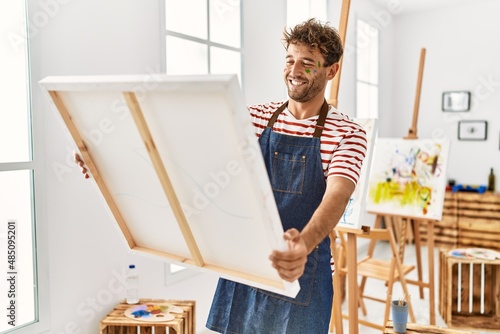 Young hispanic man smiling confident holding canvas at art studio