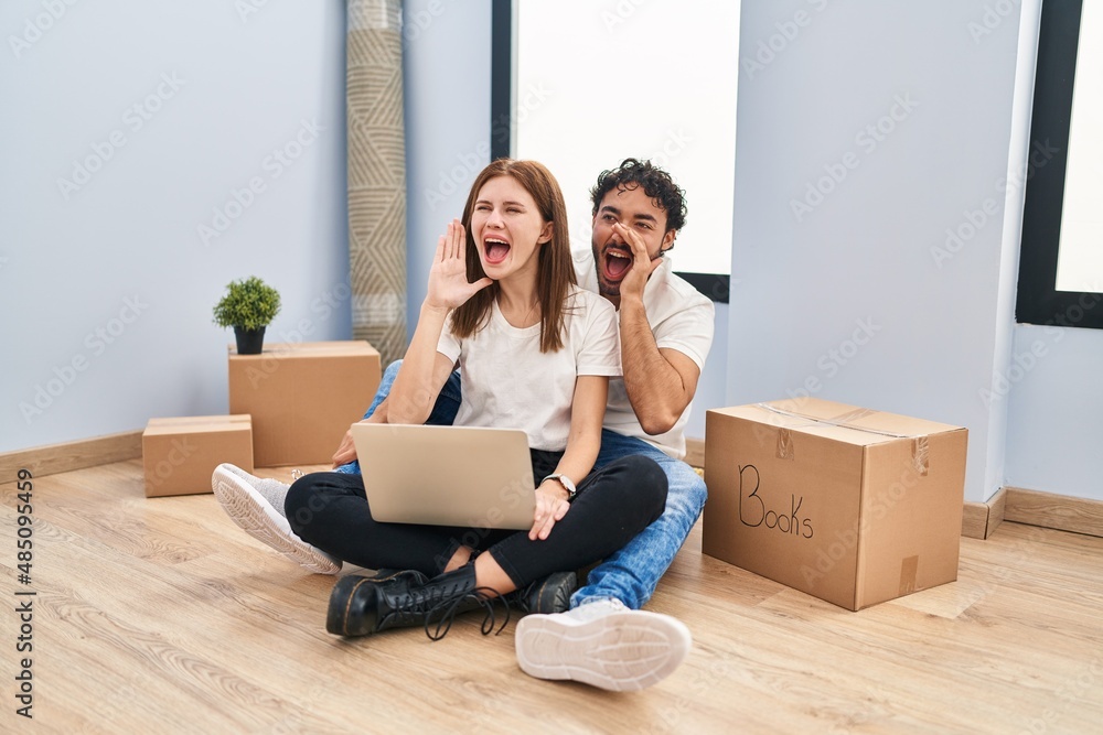 Young couple using laptop at new home shouting and screaming loud to side with hand on mouth. communication concept. Young couple using laptop at new home shouting and screaming loud to side with hand on mouth. communication concept.