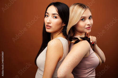 two pretty diverse girls happy posing together: blond and brunette, caucasian and asian on brown background, lifestyle people concept