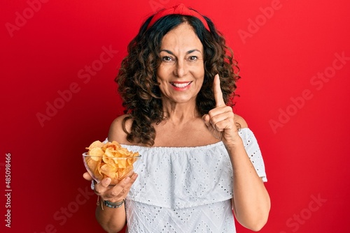 Middle age hispanic woman holding potato chip smiling with an idea or question pointing finger with happy face  number one