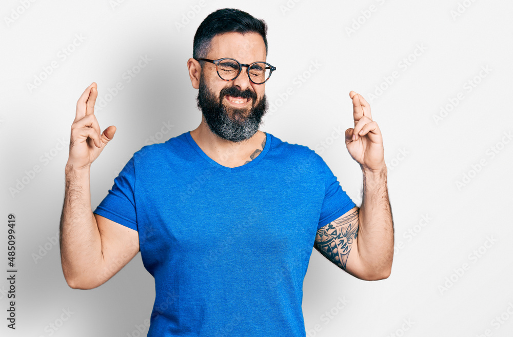 Hispanic man with beard wearing casual t shirt and glasses gesturing finger crossed smiling with hope and eyes closed. luck and superstitious concept.