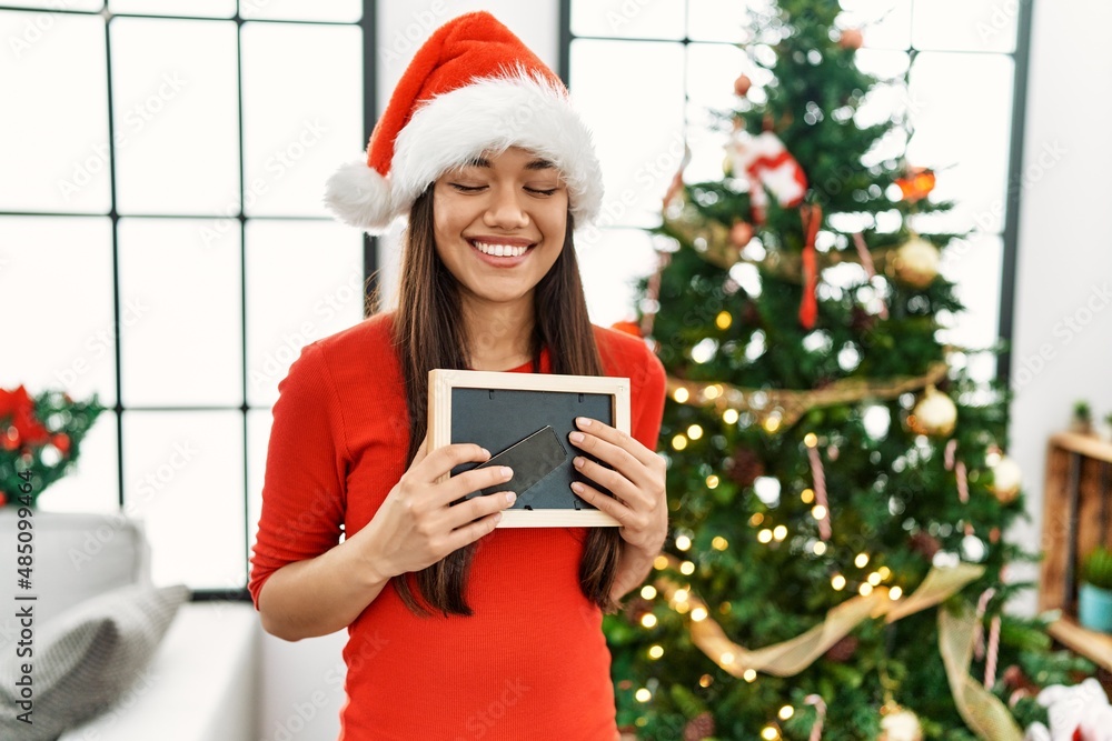 Young latin woman holding photo standing by christmas tree at home Young latin woman holding photo standing by christmas tree at home