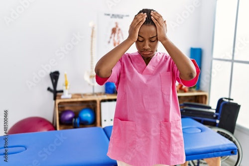 Young african american woman working at pain recovery clinic suffering from headache desperate and stressed because pain and migraine. hands on head.
