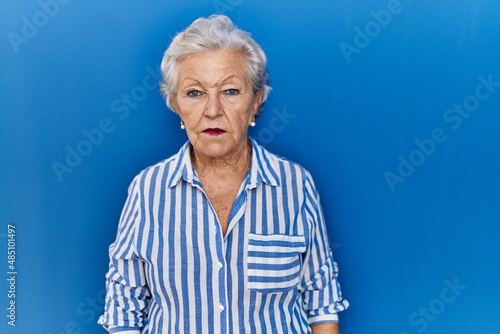 Senior woman with grey hair standing over blue background relaxed with serious expression on face. simple and natural looking at the camera.