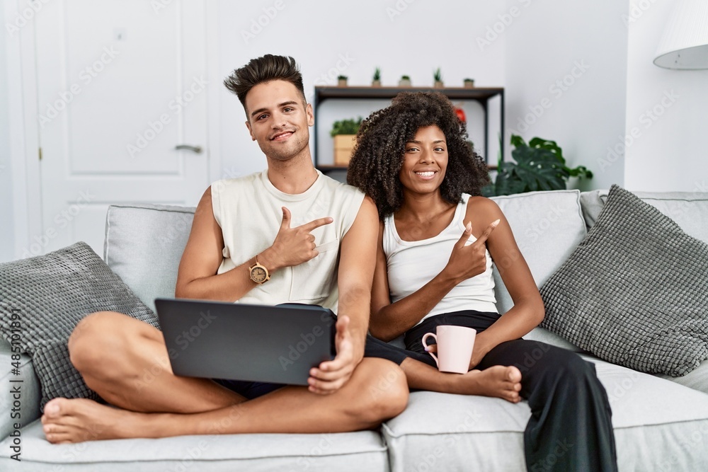 Young interracial couple using laptop at home sitting on the sofa cheerful with a smile on face pointing with hand and finger up to the side with happy and natural expression