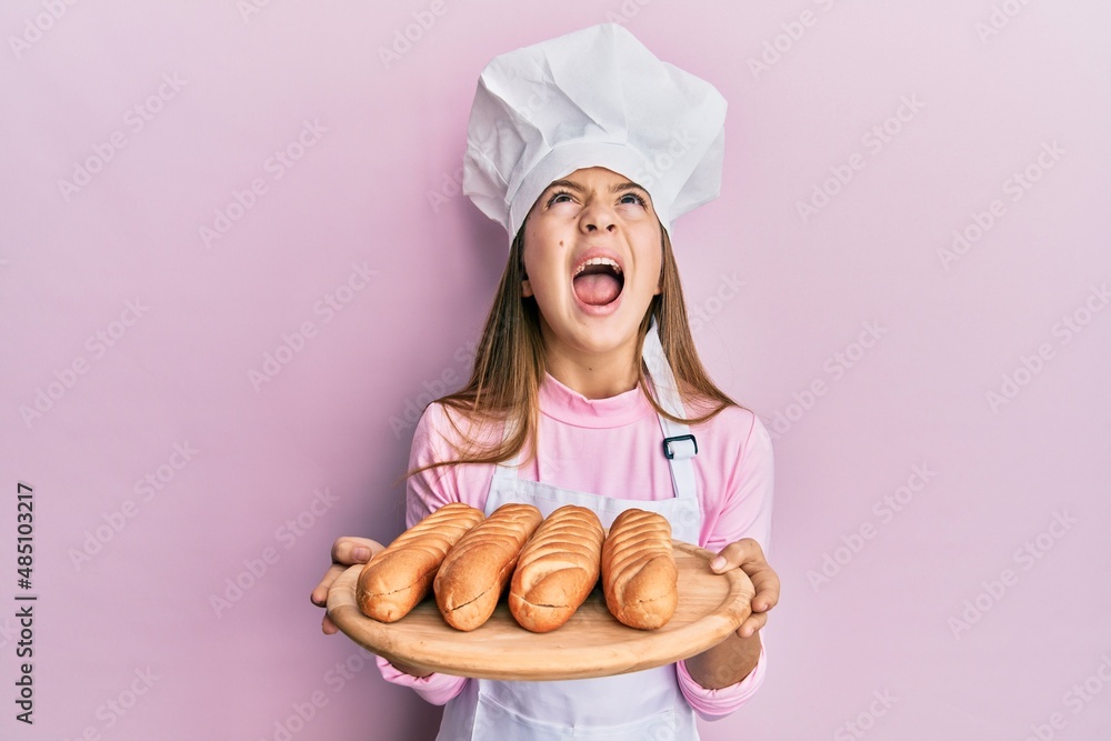 Beautiful brunette little girl wearing baker uniform holding homemade bread angry and mad screaming frustrated and furious, shouting with anger looking up. Beautiful brunette little girl wearing baker uniform holding homemade bread angry and mad screaming frustrated and furious, shouting with anger looking up.
