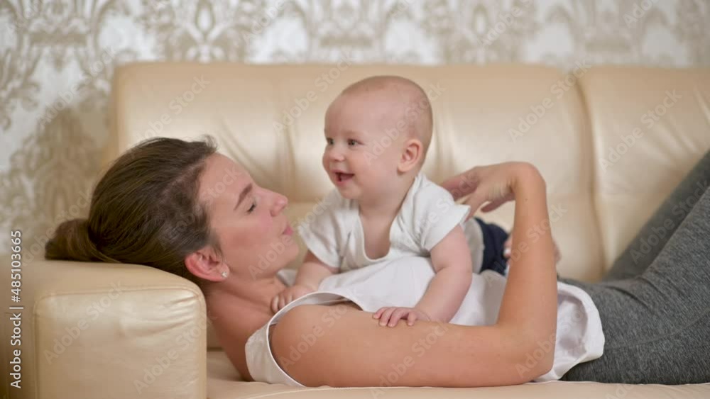 Young happy mother is playing with her newborn baby on the sofa in the morning. smiling mum enjoying bonding cuddling hugging spending time with baby. Parenthood, Childhood. Young happy mother is playing with her newborn baby on the sofa in the morning. smiling mum enjoying bonding cuddling hugging spending time with baby. Parenthood, Childhood.