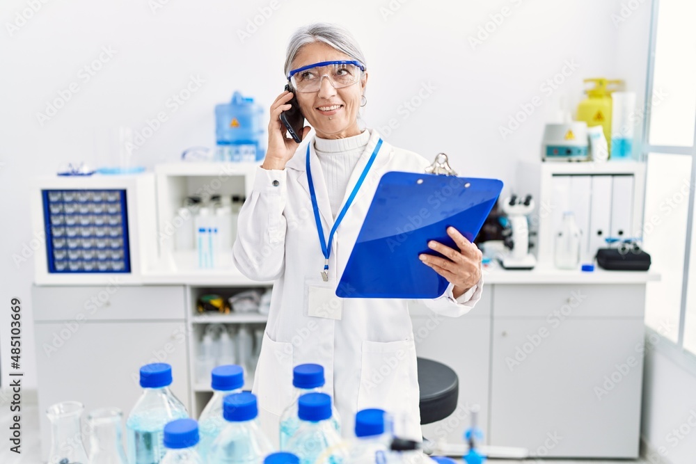Middle age grey-haired woman wearing scientist uniform talking on the smartphone at laboratory Middle age grey-haired woman wearing scientist uniform talking on the smartphone at laboratory