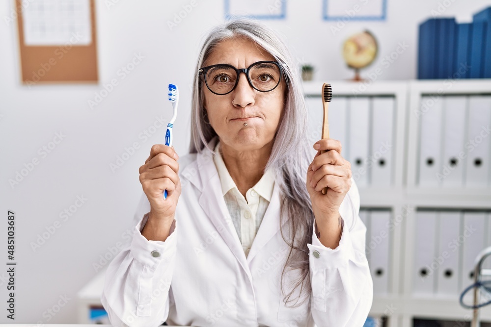 Middle age grey-haired woman working at dentist clinic holding electric and recycled teethbrush puffing cheeks with funny face. mouth inflated with air, catching air. Middle age grey-haired woman working at dentist clinic holding electric and recycled teethbrush puffing cheeks with funny face. mouth inflated with air, catching air.