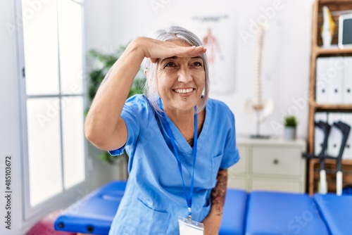 Middle age grey-haired woman wearing physiotherapist uniform at medical clinic very happy and smiling looking far away with hand over head. searching concept.