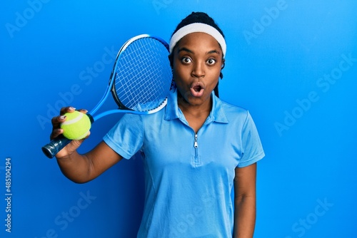 African american woman with braided hair playing tennis holding racket and ball scared and amazed with open mouth for surprise, disbelief face © Krakenimages.com