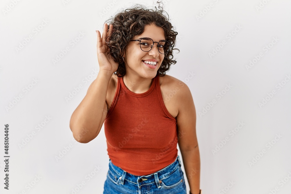 Young hispanic woman wearing glasses standing over isolated background smiling with hand over ear listening an hearing to rumor or gossip. deafness concept. Young hispanic woman wearing glasses standing over isolated background smiling with hand over ear listening an hearing to rumor or gossip. deafness concept.