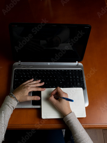 vista desde arriba de un ordenador portátil , y manos de mujer tomando notas en un cuaderno encima de un escritorio de madera. 