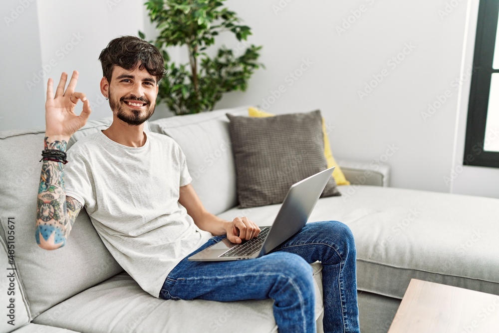 Hispanic man with beard sitting on the sofa smiling positive doing ok sign with hand and fingers. successful expression.