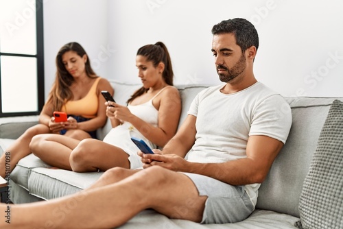 Three hispanic friends with serious expression using smartphone sitting on the sofa at home.