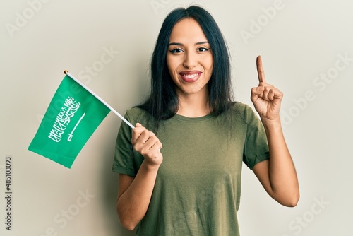 Young hispanic girl holding kingdom of saudi arabia flag smiling with an idea or question pointing finger with happy face, number one
