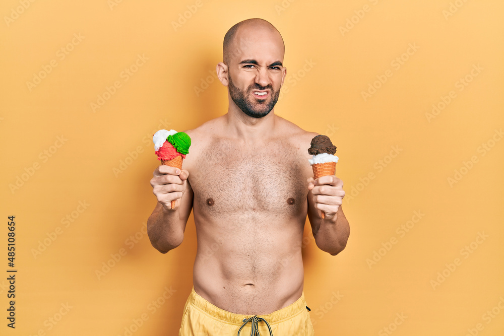 Fototapeta premium Young bald man wearing swimwear holding two ice cream cones clueless and confused expression. doubt concept.