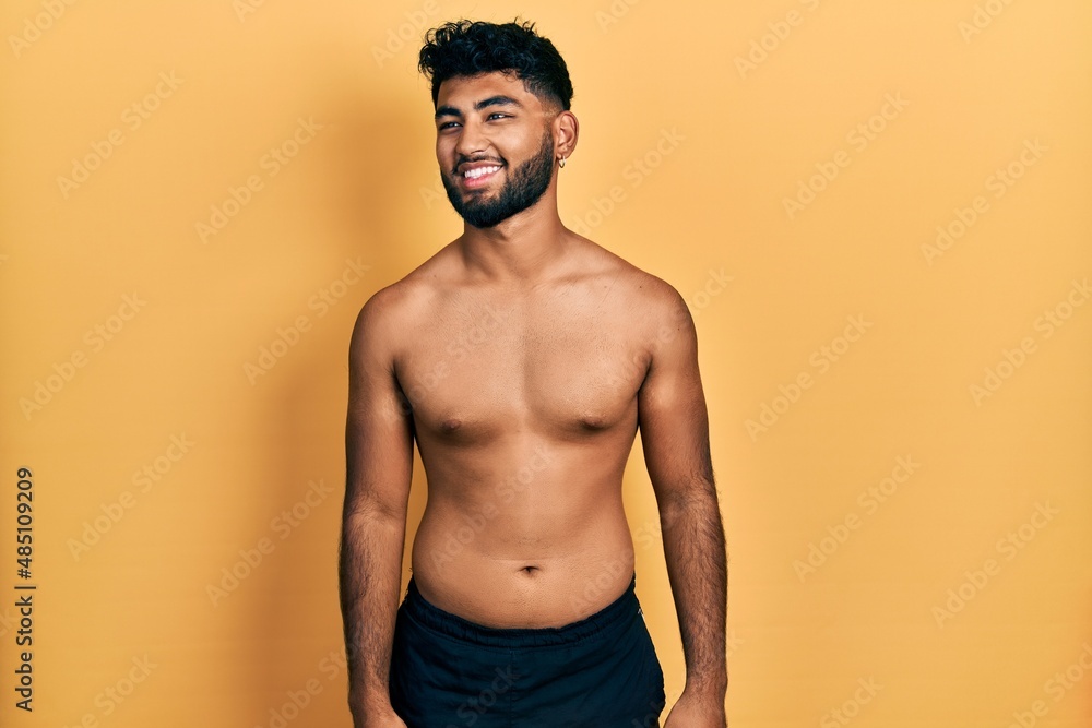 Arab man with beard wearing swimwear shirtless looking away to side with smile on face, natural expression. laughing confident.