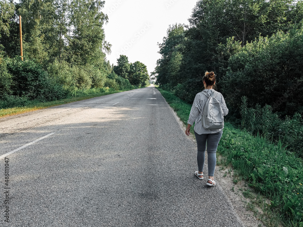 a female tourist with a backpack walks along an asphalt road in the countryside. a female tourist with a backpack walks along an asphalt road in the countryside.