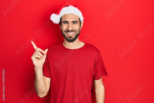 Young hispanic man wearing christmas hat with a big smile on face, pointing with hand and finger to the side looking at the camera.