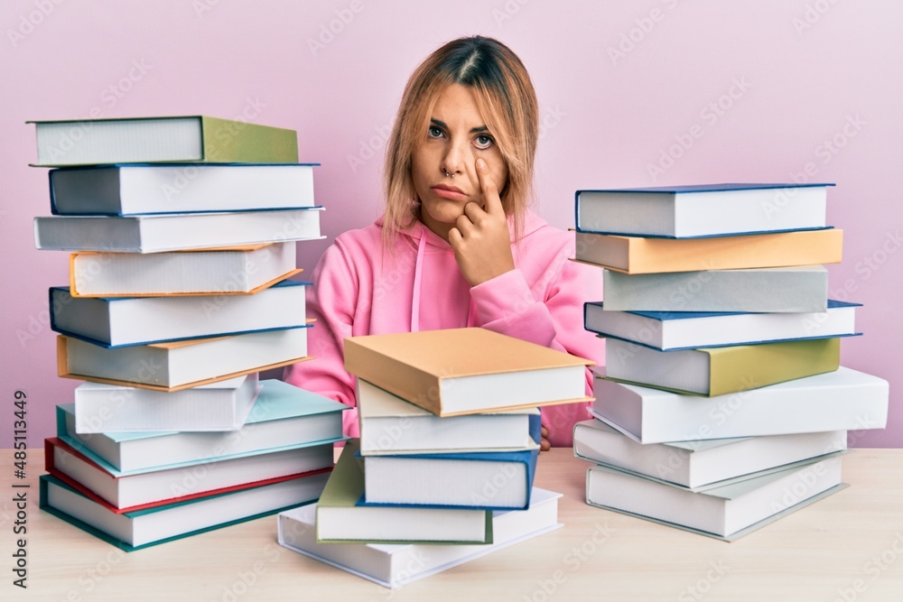 Young caucasian woman sitting on the table with books pointing to the eye watching you gesture, suspicious expression Young caucasian woman sitting on the table with books pointing to the eye watching you gesture, suspicious expression