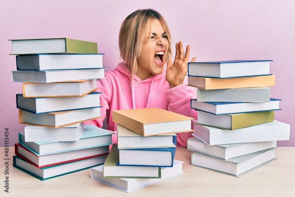 Young caucasian woman sitting on the table with books shouting and screaming loud to side with hand on mouth. communication concept. Young caucasian woman sitting on the table with books shouting and screaming loud to side with hand on mouth. communication concept.