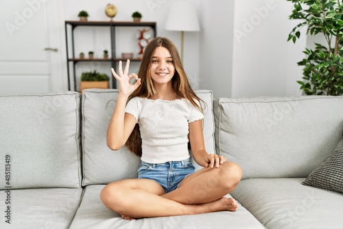 Young brunette teenager sitting on the sofa at home smiling positive doing ok sign with hand and fingers. successful expression.