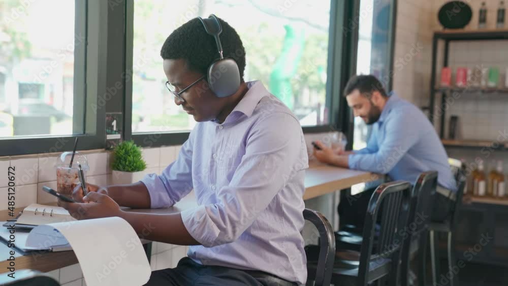 Close up of African businessman use headphone and listen music sitting in the coffee shop. Take some idea on the notebook. Looking at out side for relaxation. Working online at cafe. Business concept Close up of African businessman use headphone and listen music sitting in the coffee shop. Take some idea on the notebook. Looking at out side for relaxation. Working online at cafe. Business concept