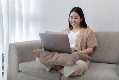 Asian woman sitting on the sofa with a laptop, Looking at laptop screen, Relaxing at home, Working through the Internet communication system, Happy lifestyle, Feel good.