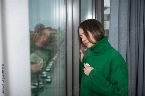 Young sick unhappy woman suffering from common cold or flu standing near window looking out wipes her nose with tissue. Self isolated ill female stay at home until she recover and get well. © Srdjan