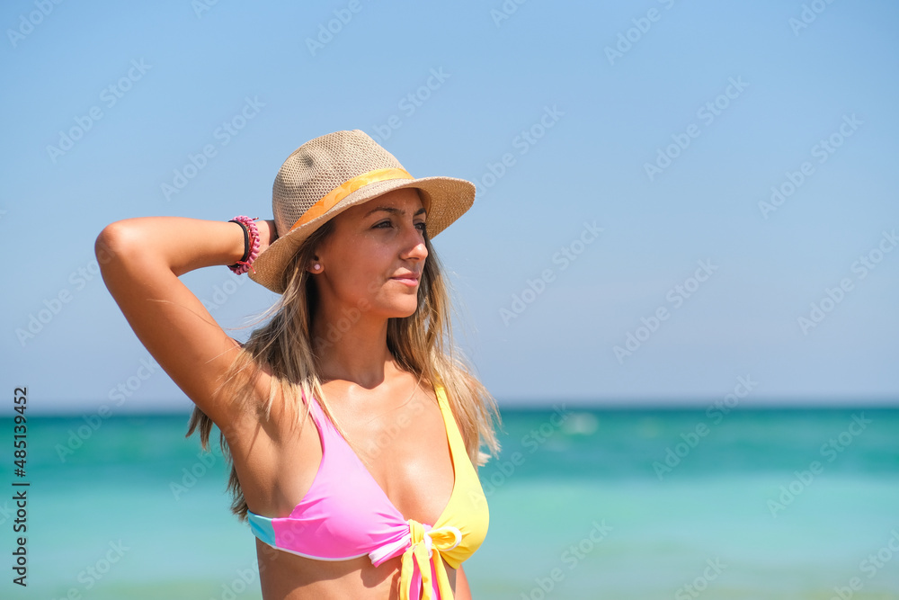 Woman in bikini on the beach with a hand on a straw hat