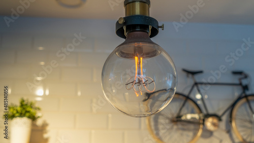 Efficient incandescent light bulb waste electricity against a tiled wall with a small bike and a plant pot. © kalyanby