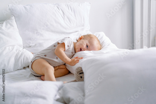 Cute toddler laying on a white bed with lots of pillows and sly smiling looking at camera.