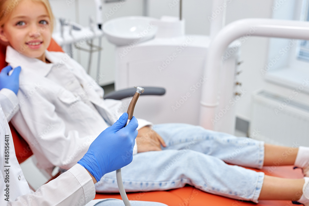 Dentist in gloves holding dental drill while treating child girl patient in modern dental office clinic, side view on adorbale excited child sitt on couch waiting for treatment. focus on dental tool Dentist in gloves holding dental drill while treating child girl patient in modern dental office clinic, side view on adorbale excited child sitt on couch waiting for treatment. focus on dental tool