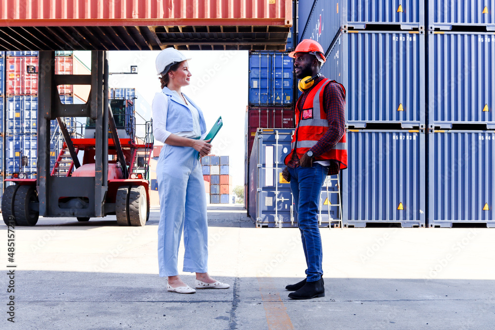 Two workers wearing safety vest and helmet discussing at logistic shipping cargo containers yard. African American engineer man talking with beautiful young woman boss with blonde hair at workplace. Two workers wearing safety vest and helmet discussing at logistic shipping cargo containers yard. African American engineer man talking with beautiful young woman boss with blonde hair at workplace.