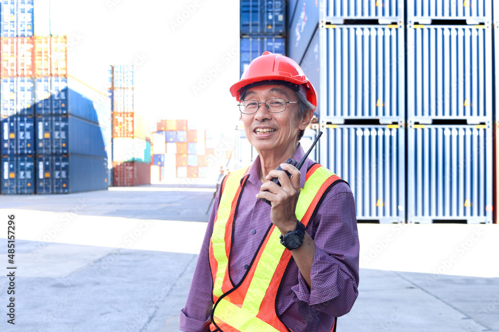 A senior elderly Asian worker engineer wearing safety vest and helmet standing and holding radio walkies talkie at logistic shipping cargo containers yard. elderly people at workplace concept. A senior elderly Asian worker engineer wearing safety vest and helmet standing and holding radio walkies talkie at logistic shipping cargo containers yard. elderly people at workplace concept.