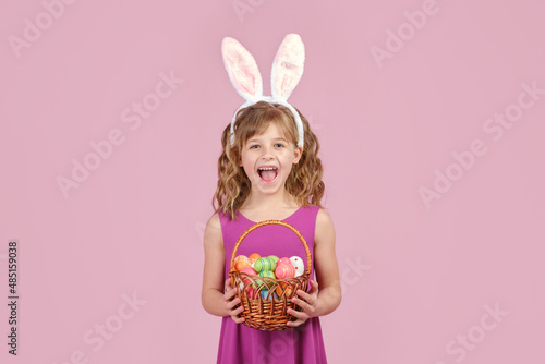 Little beautiful smiling girl in Easter bunny ears holds in hands a basket with Easter eggs on pink background 