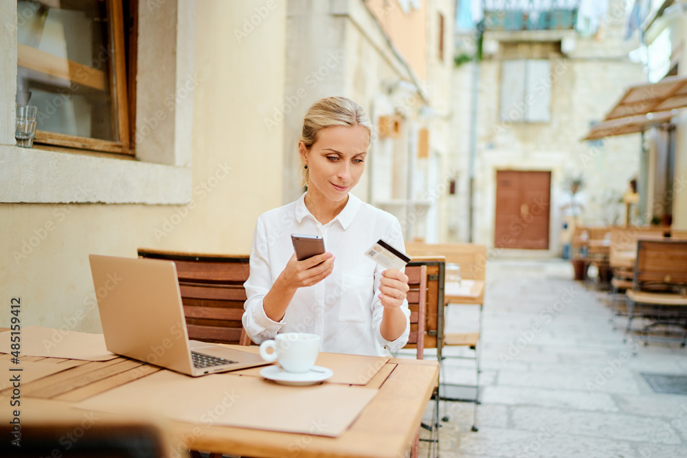 Working, traveling and online banking. Pay the bill. Pretty young woman holding plastic credit card using smartphone and laptop in sidewalk cafe on ancient europian street. Working, traveling and online banking. Pay the bill. Pretty young woman holding plastic credit card using smartphone and laptop in sidewalk cafe on ancient europian street.