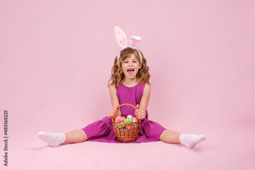 Little beautiful smiling girl in Easter bunny ears holds in hands a basket with Easter eggs and sitting on the floor on pink background 