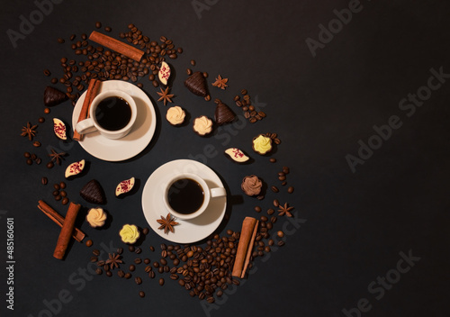 Two white cups of coffee, coffee beans, spices and chocolate candies on dark background.
