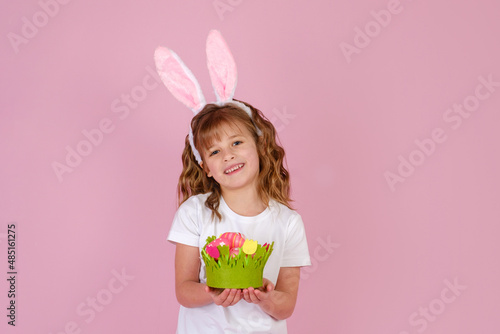 Little beautiful smiling girl in Easter bunny ears headband holds in hands Easter eggs on pink background 