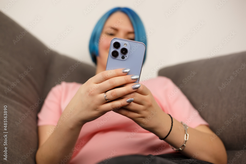 Young woman using mobile phone with triple camera. Creative white female with dyed blue hair browsing internet on modern smartphone while sitting on couch at home during lockdown. Person communicating Young woman using mobile phone with triple camera. Creative white female with dyed blue hair browsing internet on modern smartphone while sitting on couch at home during lockdown. Person communicating